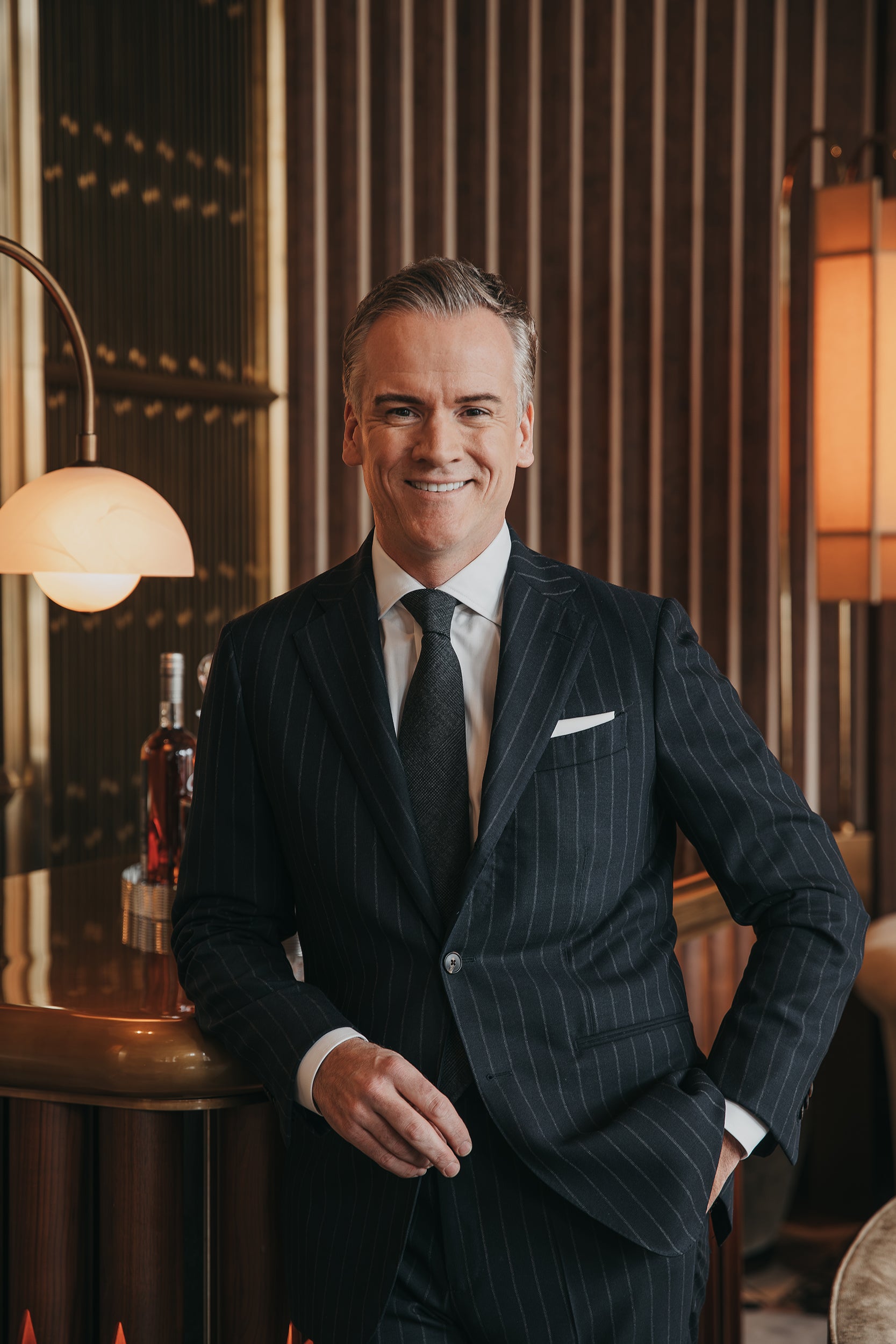 Man in a suit standing in an elegant interior setting with warm lighting.