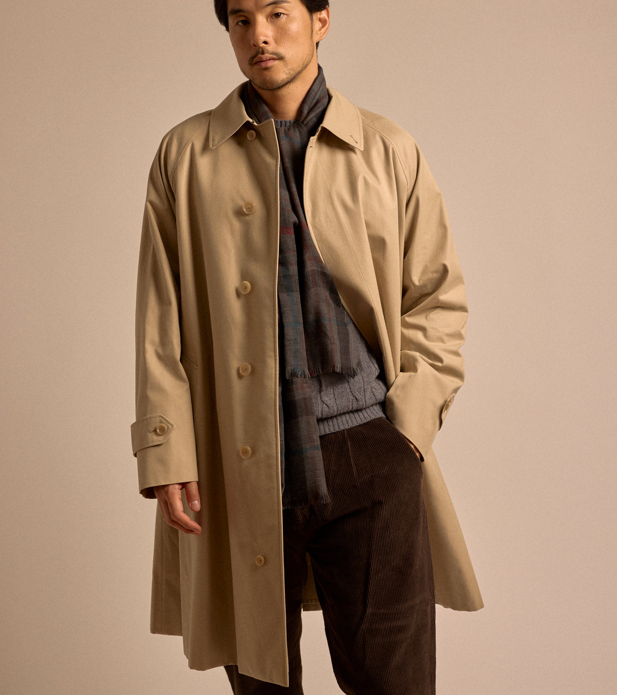 Model wearing Sanyo 100 Year Lightweight Gabardine Balmacaan Coat in Beige. Styled with grey scarf, grey cable knit sweater and brown corduroy trousers.