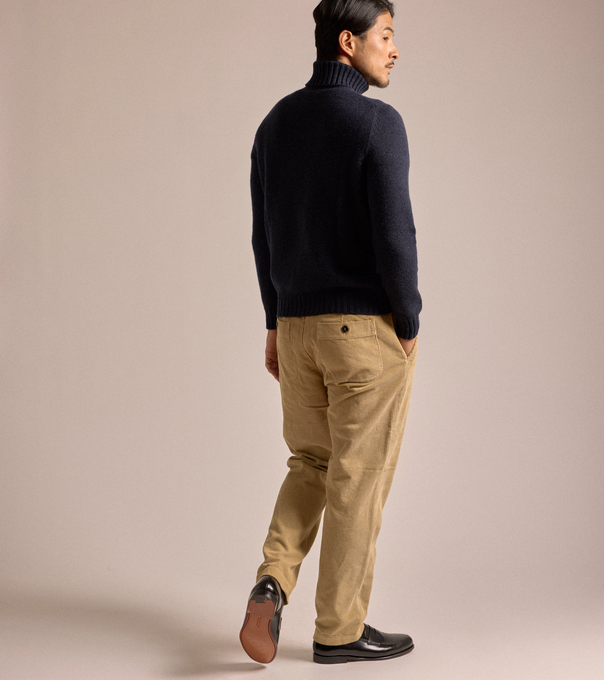 Model wearing the East Harbour Surplus Cord Oxnard Trousers in Fawn, shown from the back and styled with a navy roll neck sweater and leather shoes.