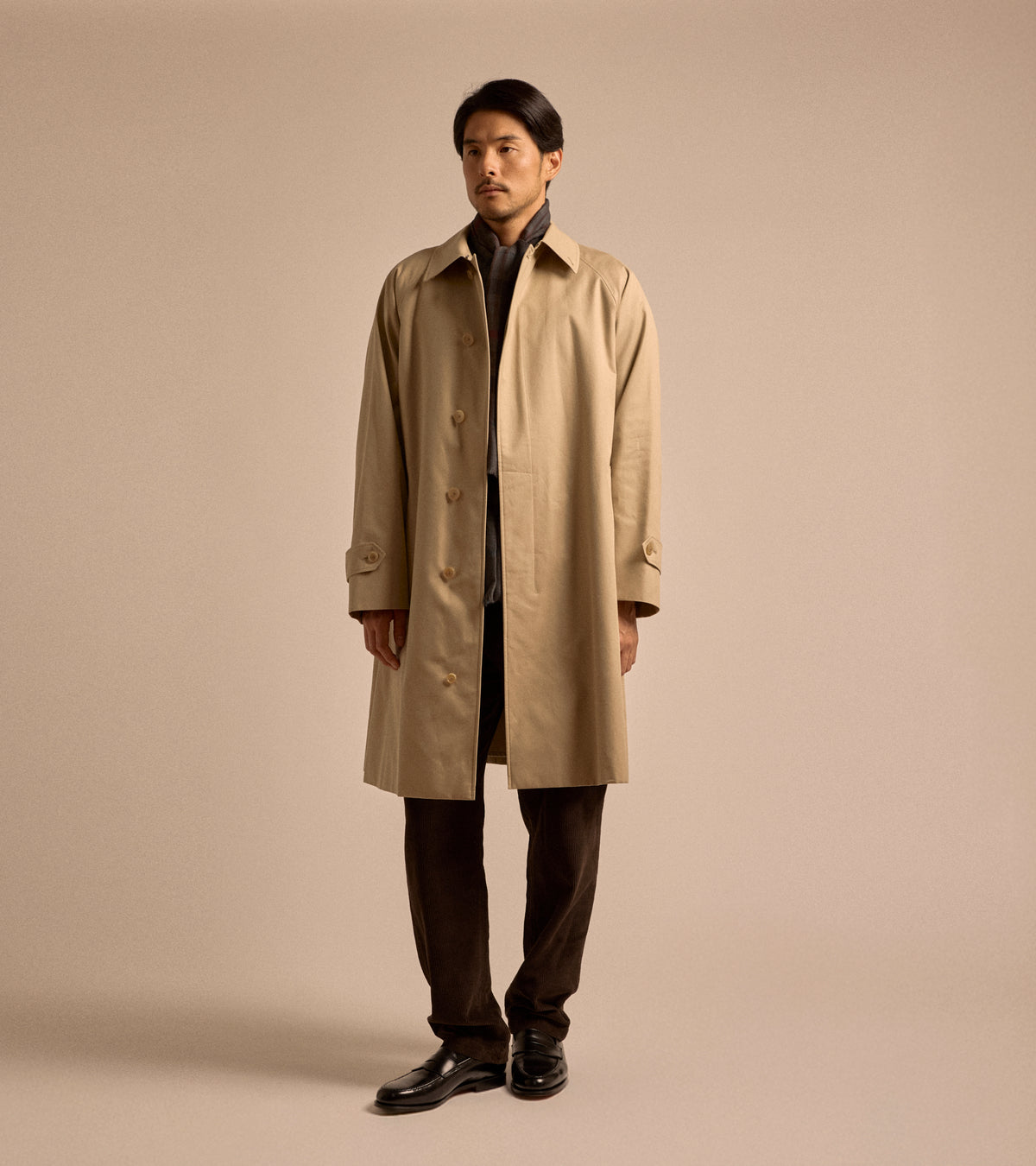 Model wearing the Sanyo 100 Year Lightweight Gabardine Balmacaan Coat in Beige. Shown here in full length, styled with grey scarf, brown corduroy trousers and black leather loafers.