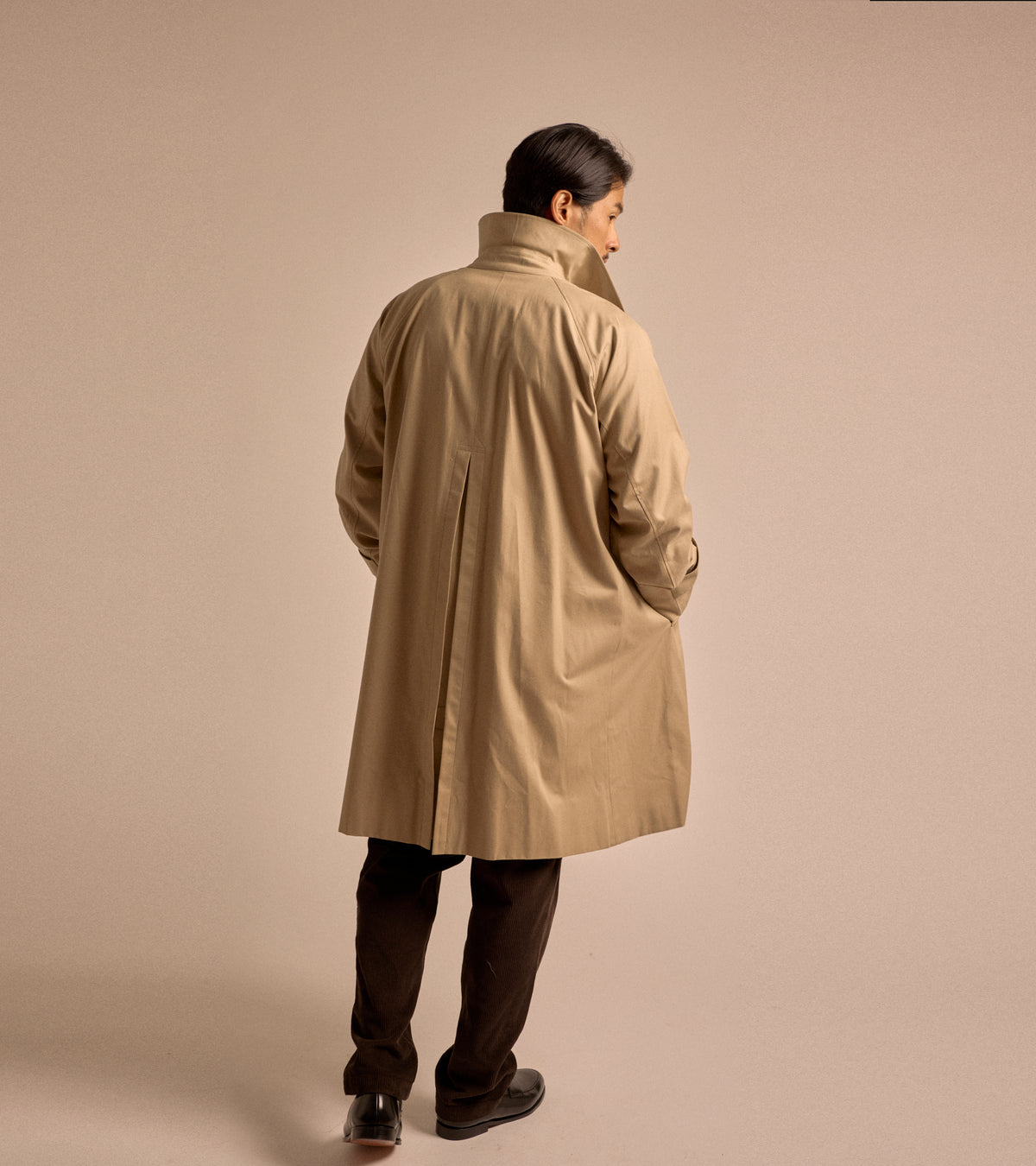 Model is wearing the Sanyo 100 Year Lightweight Gabardine Balmacaan Coat in Beige, back view. Styled with brown trousers and black leather loafers.