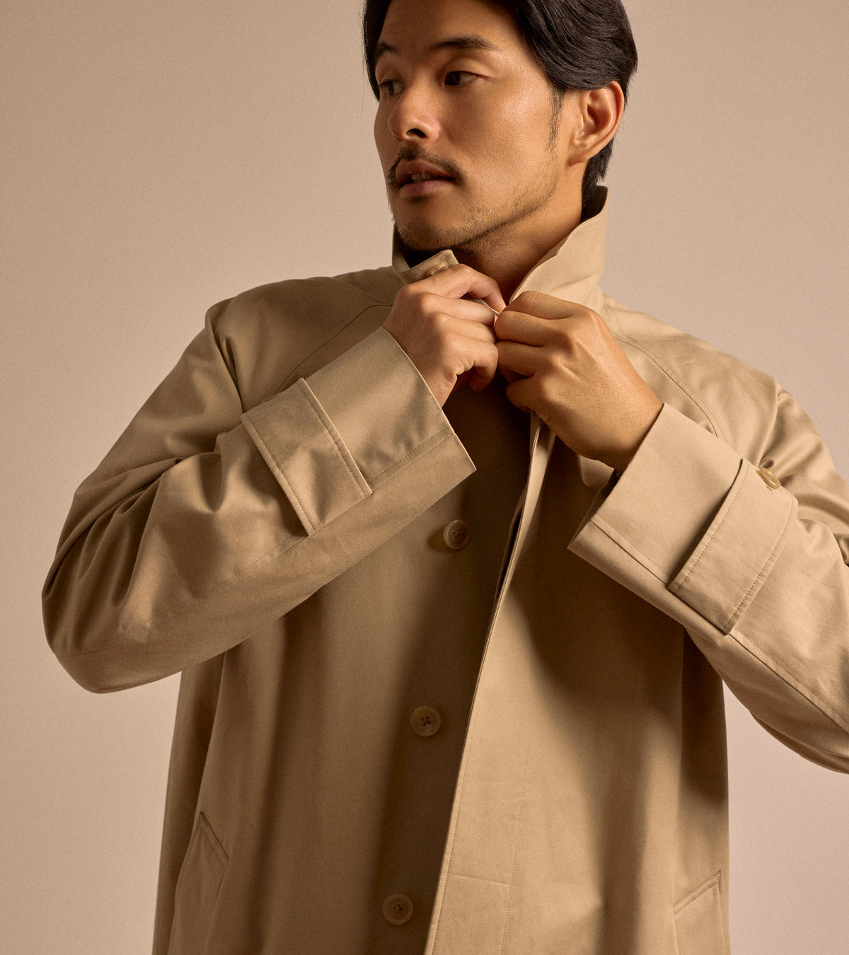 Model wearing the Sanyo 100 Year Lightweight Gabardine Balmacaan Coat in Beige. Close-up shot of the model buttoning up the collar.